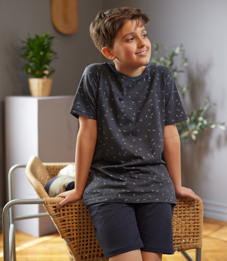 Dečak u sivoj pidžami na kratak rukavi sorcu stoji naslonjen a stolicu