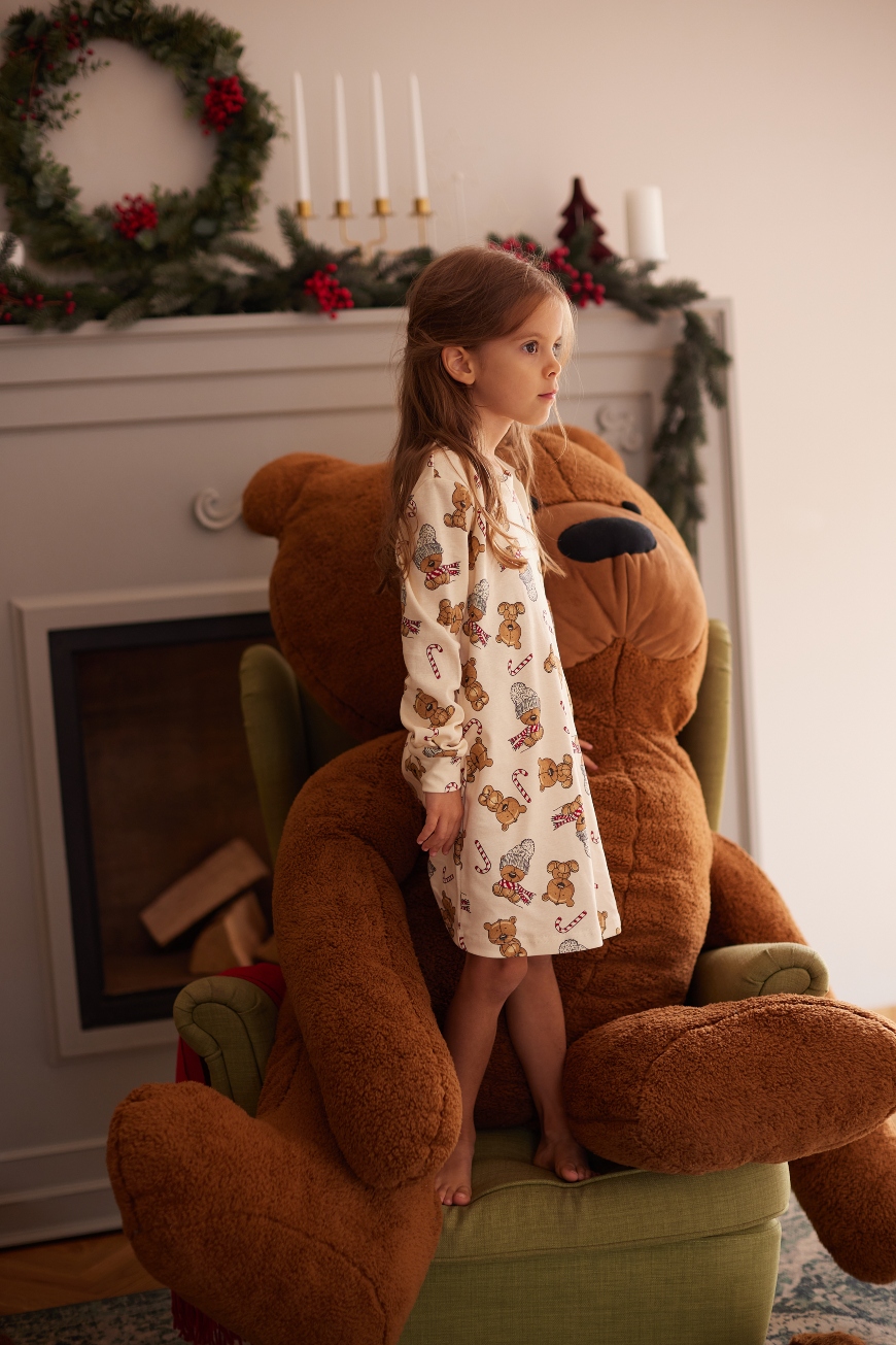 Devojčica u beloj spavaćici na dug rukav sa medvedićima stoji na fotelji sa velikim plišanim medvedom