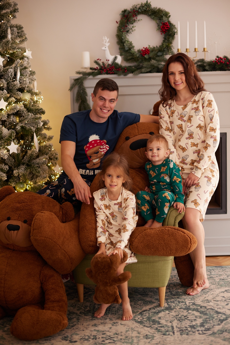 Žena i muškarac sa decom u novogodišnjim pidžamama sede na fotelji ispred jelkesa velikim plišanim medvedima