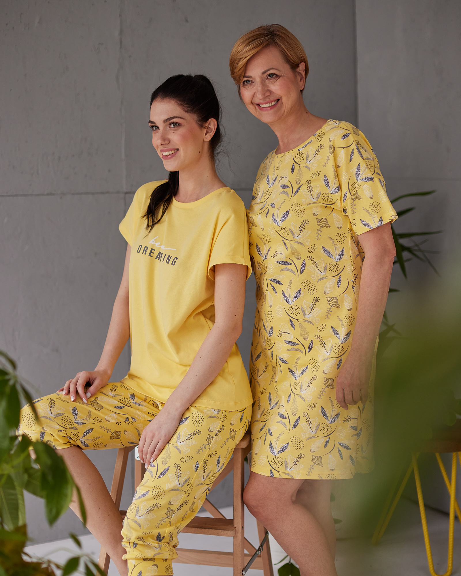 Devojka sedi i žena pored nje stoji u žutim pidžamama sa šarom listova i cveca